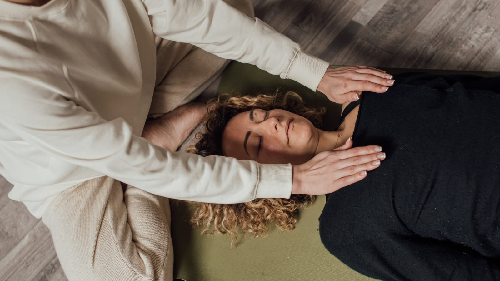 A pair of hands hovering above a still figure on a soft blanket, warm low light, the visual register of a quiet, attentive session.